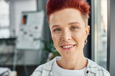 Porträt einer fröhlichen, stilvollen, queeren Person mit Piercing im Gesicht, die in die Kamera im modernen Büro blickt