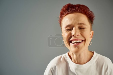 Porträt einer rothaarigen bigeschlechtlichen Person mit Piercing im Gesicht, die mit geschlossenen Augen vor grauem Hintergrund lacht