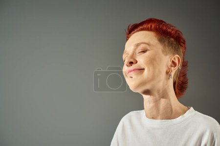 portrait of redhead bigender person with facial piercing smiling with closed eyes on grey backdrop