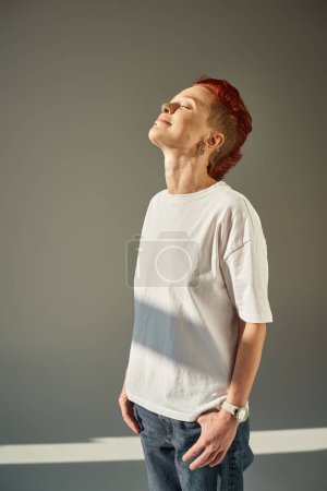 Foto de Persona queer feliz y única en camiseta blanca y jeans de pie a la luz del sol sobre fondo gris - Imagen libre de derechos