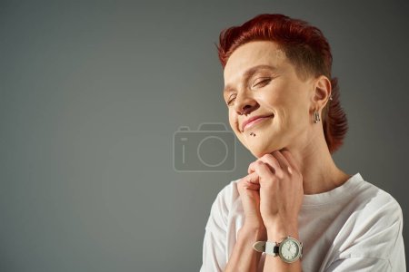 Photo for Portrait of redhead bigender person with facial piercing smiling with closed eyes on grey backdrop - Royalty Free Image