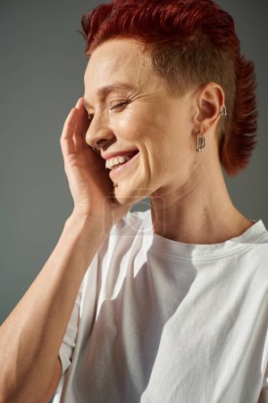 redhead bigender person with piercing smiling with closed eyes and hand near face on grey, portrait