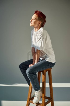 Photo for Queer person in white t-shirt and jeans smiling with closed eyes on stool in sunlight on grey - Royalty Free Image