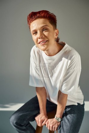 Photo for Smiling bigender person in white t-shirt sitting on stool in sunlight and looking at camera on grey - Royalty Free Image