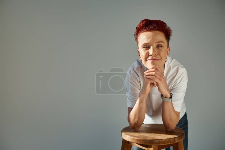 Photo for Positive bigender person in white t-shirt posing with stool in sunlight looking at camera on grey - Royalty Free Image