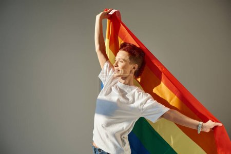 Photo for Happy and unique queer person in white t-shirt posing with rainbow colors LGBT flag on grey backdrop - Royalty Free Image