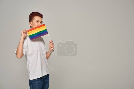 redhead queer model in white t-shirt posing with small LGBT flat near face looking at camera on grey