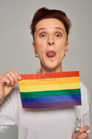 rothaariges queeres Model mit Piercing und kleiner LGBT-Wohnung, die hervorsteht und in die Kamera auf grau schaut