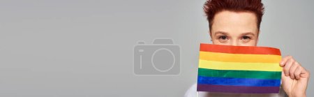 joyful redhead queer person obscuring face with small LGBT flag looking at camera on grey, banner