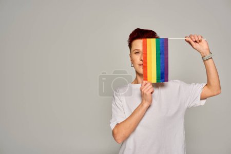 Photo for Redhead queer person in white t-shirt obscuring face with small LGBT flag looking at camera on grey - Royalty Free Image