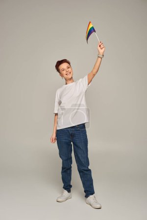 cheerful queer person in white t-shirt holding small LGBT flag in raised hand while standing on grey