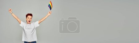 excited queer person in white t-shirt standing with small LGBT flag and screaming on grey, banner