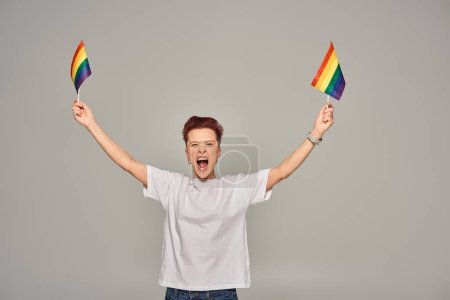 Photo for Excited queer person in white t-shirt standing with small LGBT flag and screaming on grey backdrop - Royalty Free Image