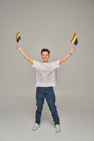 Foto de Alegre persona queer en camiseta blanca y jeans posando con pequeñas banderas LGBT en manos levantadas sobre gris - Imagen libre de derechos