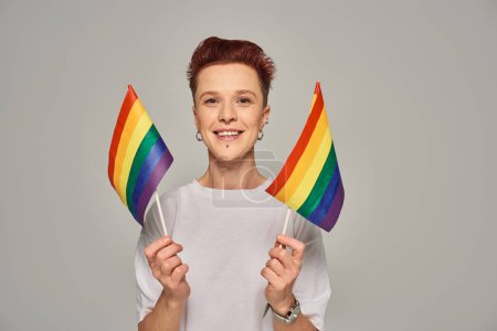 joyful queer person in white t-shirt holding small LGBT flags and looking at camera on grey backdrop