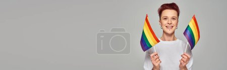 Foto de Alegre persona queer en camiseta blanca sosteniendo pequeñas banderas LGBT y mirando a la cámara en gris, pancarta - Imagen libre de derechos