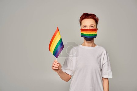 rothaarige queere Person in weißem T-Shirt und Regenbogenfarben medizinische Maske mit kleiner LGBT-Flagge auf grau
