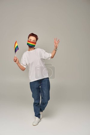 volle Länge der rothaarigen queeren Person in Regenbogenfarben medizinische Maske mit kleiner LGBT-Flagge auf grau