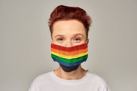 retrato de pelirroja queer persona en colores arco iris máscara médica mirando a la cámara en el fondo gris