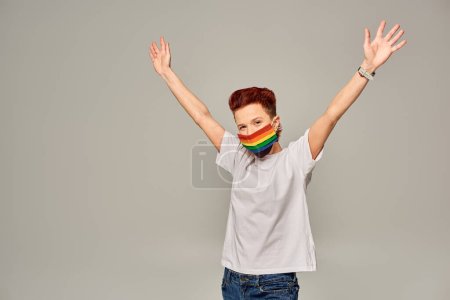 Foto de Modelo queer pelirrojo en camiseta blanca y máscara médica de colores arco iris con las manos levantadas en gris - Imagen libre de derechos