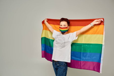 redhead queer model in white t-shirt and rainbow colors medical mask holding LGBT flag on grey