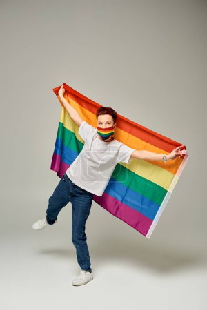 longitud completa de pelirroja modelo queer en colores arco iris máscara médica sosteniendo bandera LGBT en gris