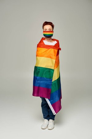 longitud completa de pelirroja queer persona en arco iris colores máscara médica posando en la bandera LGBT en gris