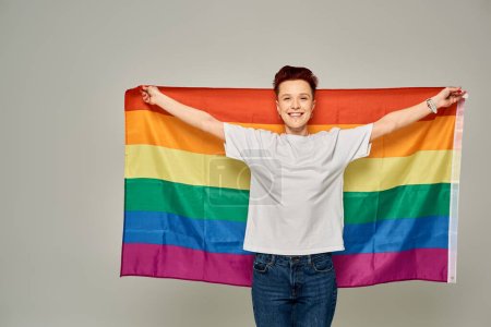 Foto de Alegre pelirroja persona grande en camiseta blanca de pie con bandera LGBT sobre fondo gris - Imagen libre de derechos
