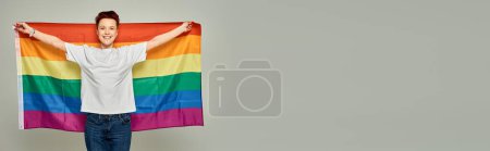 gaie rousse bigender personne en t-shirt blanc debout avec drapeau LGBT sur fond gris, bannière