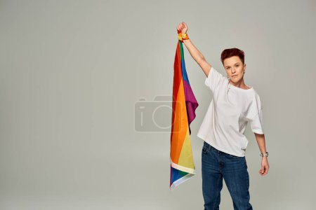 ernste rothaarige bigender Person in weißem T-Shirt und Jeans mit LGBT-Flagge auf grauem Hintergrund
