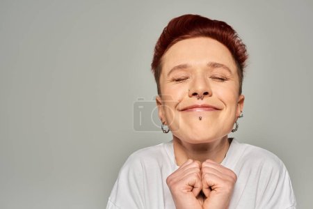 excited redhead bigender person with facial piercing smiling with closed eyes on grey, emotions