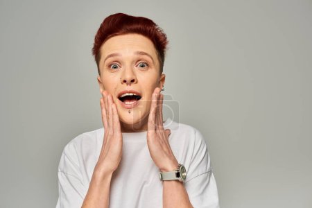 amazed redhead queer person with open mouth touching face and looking at camera on grey backdrop
