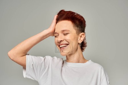 Photo for Cheerful non-binary person in white t-shirt touching red hair and smiling with closed eyes on grey - Royalty Free Image