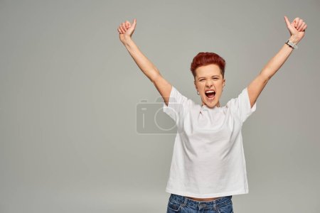 excited non-binary person in white t-shirt showing thumbs up with raised hands and screaming on grey