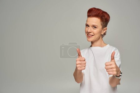 glückliche rothaarige nichtbinäre Person in weißem T-Shirt, die Daumen zeigt und in die Kamera lächelt auf grau