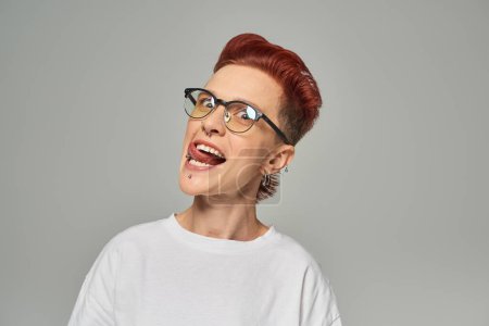 Photo for Funny and cheerful non-binary person in white t-shirt and eyeglasses sticking out tongue on grey - Royalty Free Image