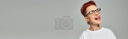 Photo for Funny and cheerful queer person in white t-shirt and eyeglasses sticking out tongue on grey, banner - Royalty Free Image