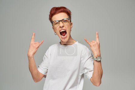 excited redhead bigender person in eyeglasses showing rock sign with hands and screaming on grey