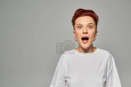 impressed bigender person in white t-shirt standing with open mouth and looking at camera on grey