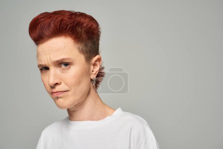 Photo for Offended and upset redhead non-binary person in white t-shirt looking at camera on grey backdrop - Royalty Free Image