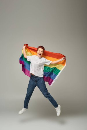 Photo for Overjoyed queer person in white t-shirt and jeans jumping and levitating with LGBT flag on grey - Royalty Free Image