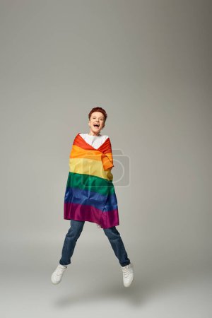 Foto de Persona queer muy alegre en camiseta blanca y pantalones vaqueros saltando y levitando con bandera LGBT en gris - Imagen libre de derechos