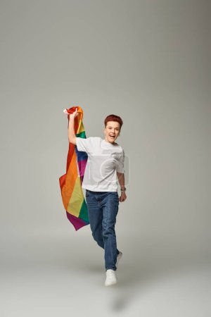 Foto de Persona queer llena de alegría en camiseta blanca y jeans y levitando con bandera LGBT sobre fondo gris - Imagen libre de derechos