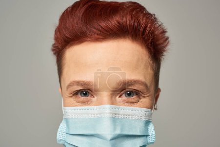 close up portrait of redhead queer person with happy gaze in medical mask looking at camera on grey