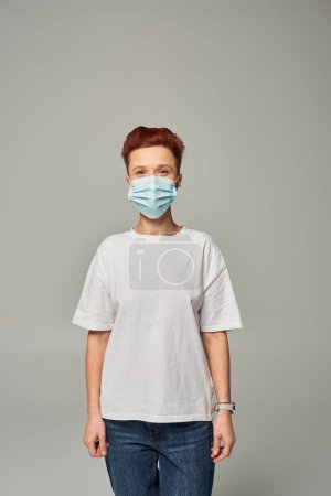 positive redhead queer person win white t-shirt and medical mask standing on grey backdrop