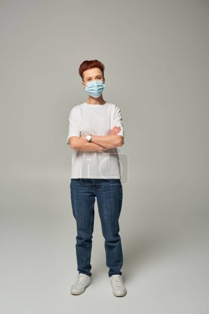 positive redhead queer person win white t-shirt and medical mask standing with folded arms on grey