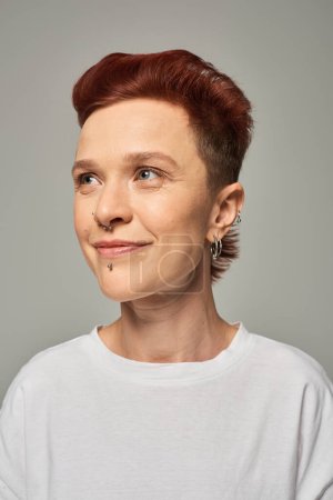 portrait of redhead non-binary model with facial piercing smiling and looking away on grey