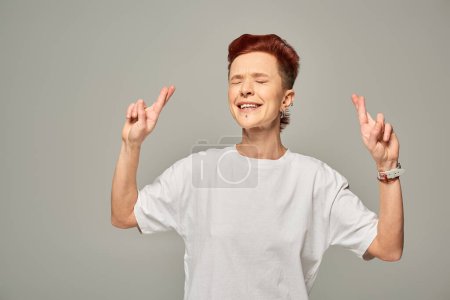angespannte bigeschlechtliche Person im weißen T-Shirt drückt dem Glück mit geschlossenen Augen auf grau stehend die Daumen