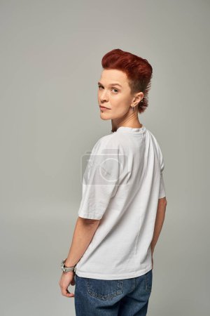 confident and serious redhead non-binary person in white t-shirt looking at camera on grey