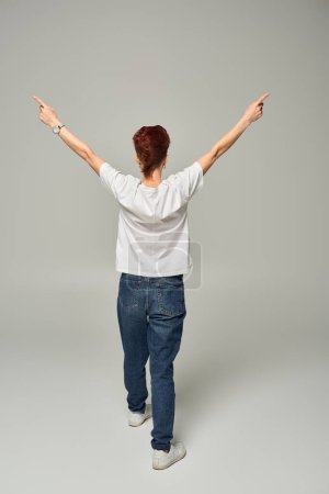 back view of redhead queer person in white t-shirt and jeans pointing aside with fingers on grey